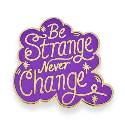 Purple and gold enamel pin with 'Be Strange Never Change' text on a white background