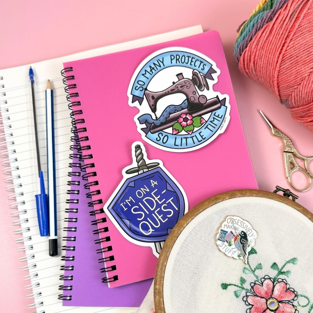 Stickers with sewing-themed designs on a pink notebook with embroidery hoop and yarn.