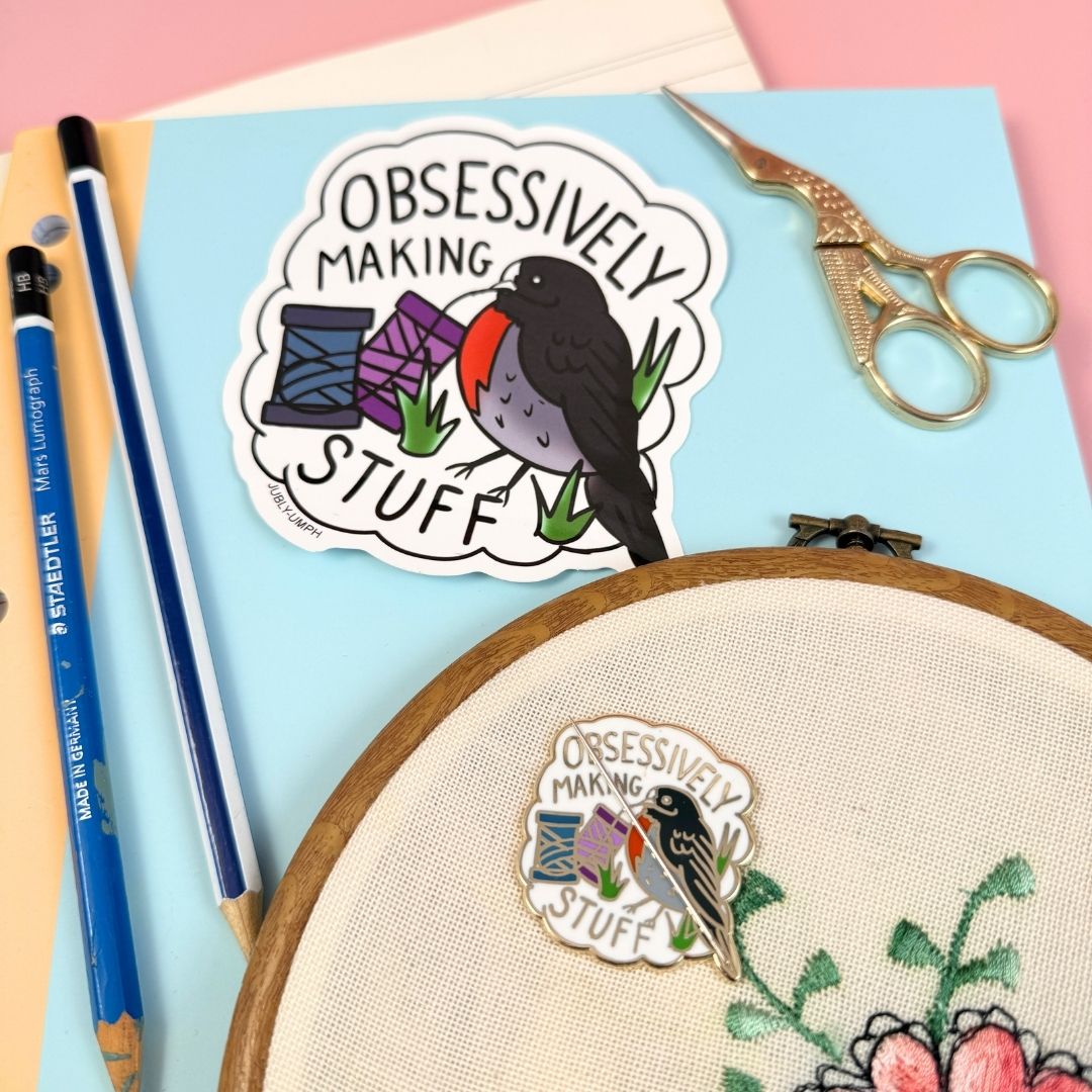 Sticker with a bird design and text 'Obsessively Making Stuff' on a blue background with embroidery hoop and tools.
