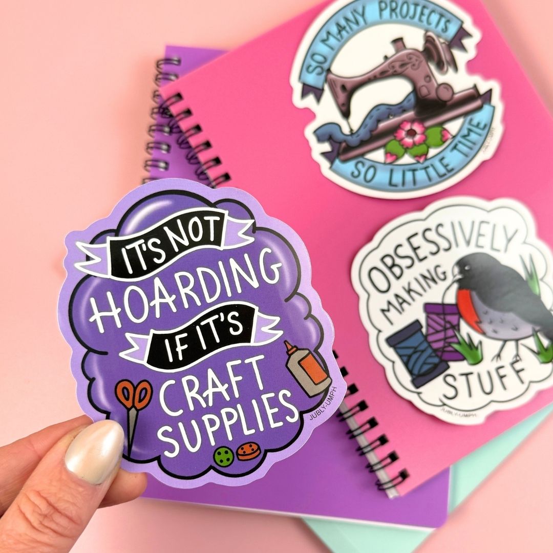 Hand holding a purple sticker with text about craft supplies on a pink background with other stickers.